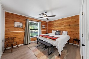 Un dormitorio con cama, un banco, ventilador de techo, paredes de madera, una ventana y una puerta.