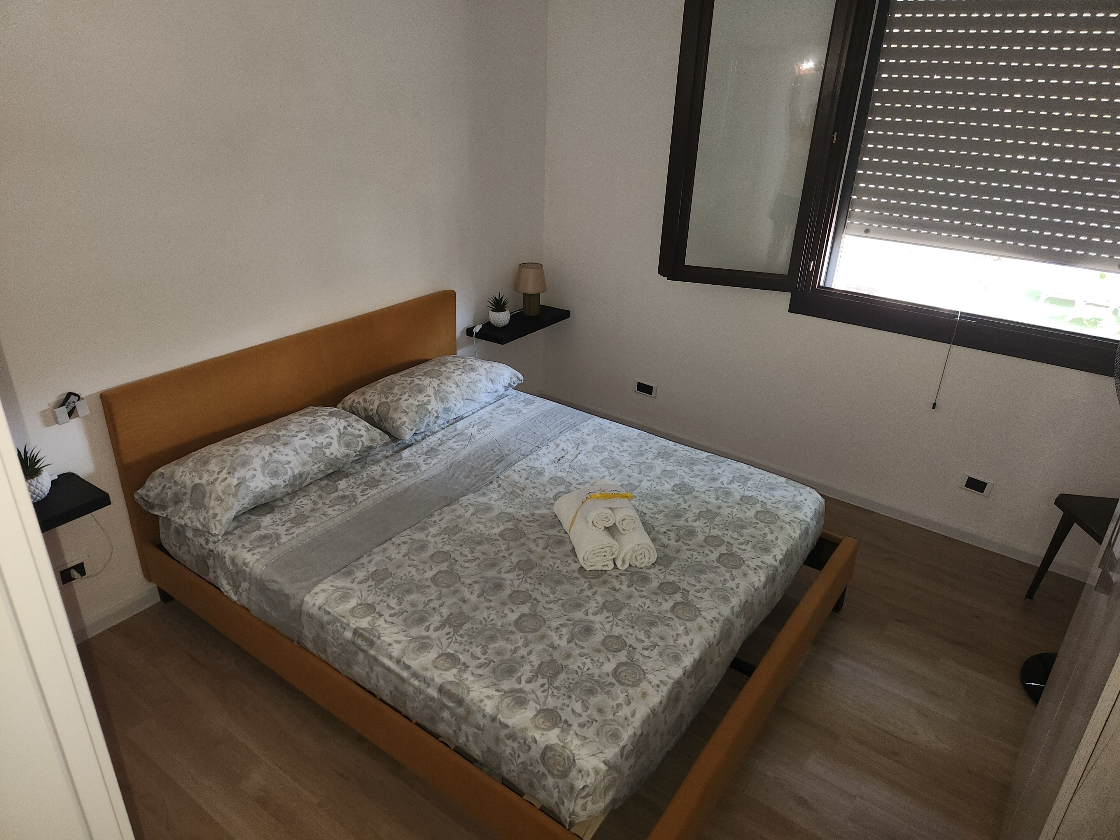 Ein Einzelbett mit Holz-Kopfteil, ein Nachttisch mit Lampe und ein Fenster mit Jalousien.