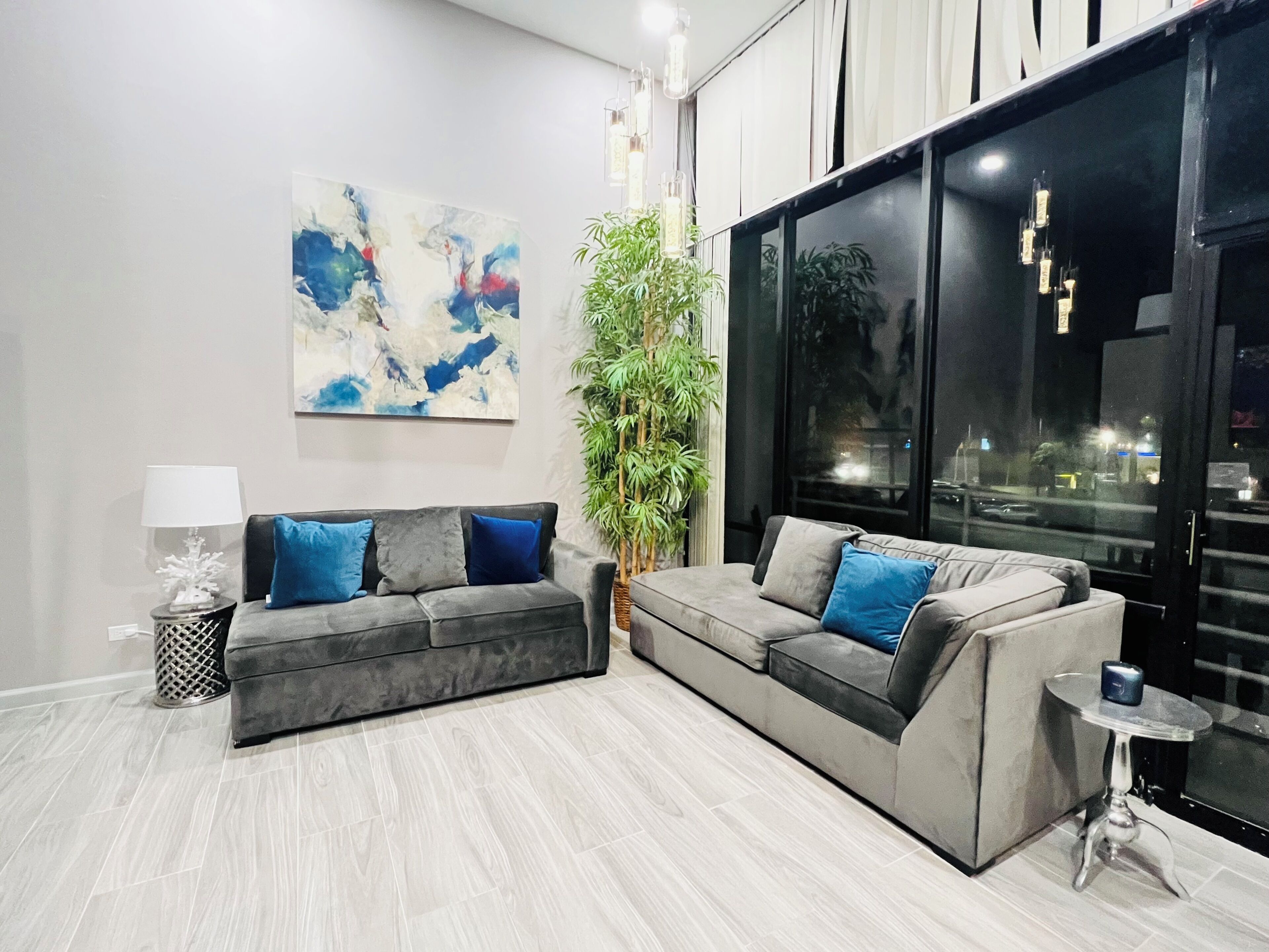 A modern living room with a grey sofa set, a large abstract painting, a potted plant, and a view through a large window.