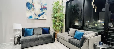 A modern living room with a grey sofa set, a large abstract painting, a potted plant, and a view through a large window.