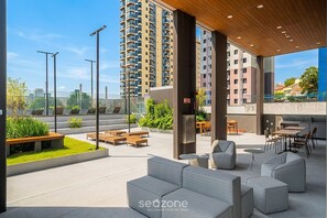 Ein moderner Außenbereich mit Sofas und Tischen, umgeben von viel Grün und Blick auf hohe Gebäude.