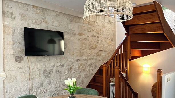 A dining area with a round wooden table, green upholstered chairs, a vase with white flowers, a stone wall, a flat-screen TV, and a staircase with wooden railings.