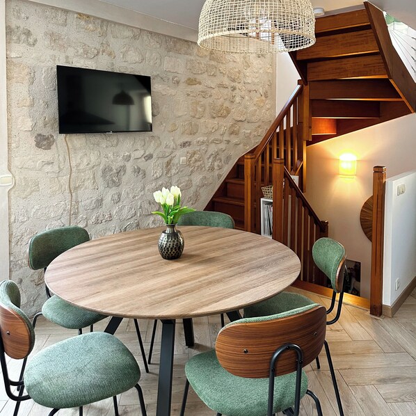 A dining area with a round wooden table, green upholstered chairs, a vase with white flowers, a stone wall, a flat-screen TV, and a staircase with wooden railings.