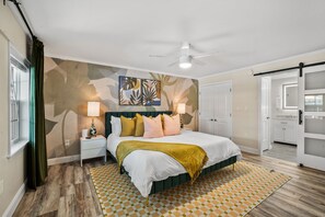 A bedroom with a bed, nightstands, a ceiling fan, a patterned rug, and a door leading to another room.
