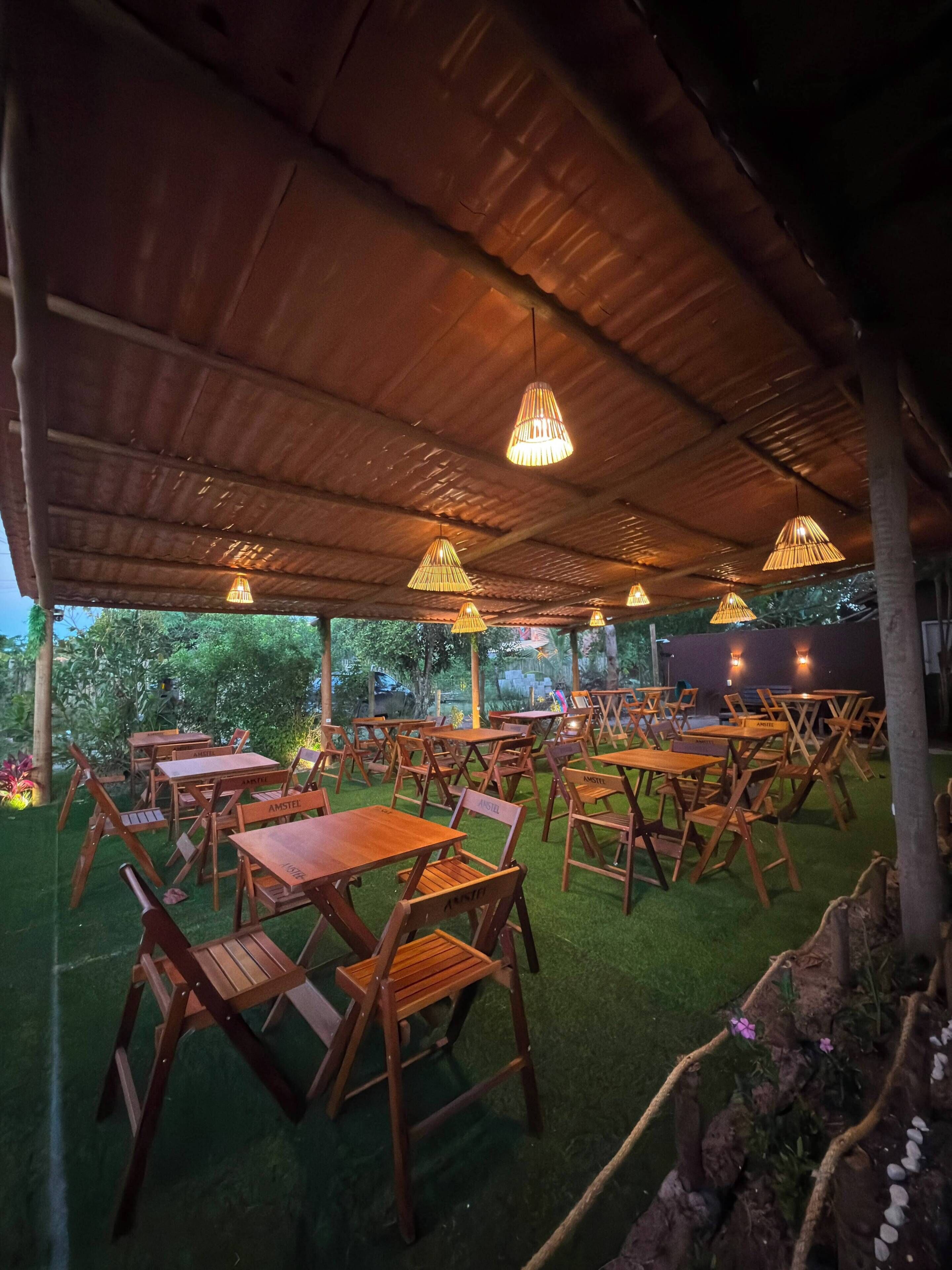 Un área de comedor al aire libre con mesas y sillas bajo un techo de madera, rodeada de follaje.