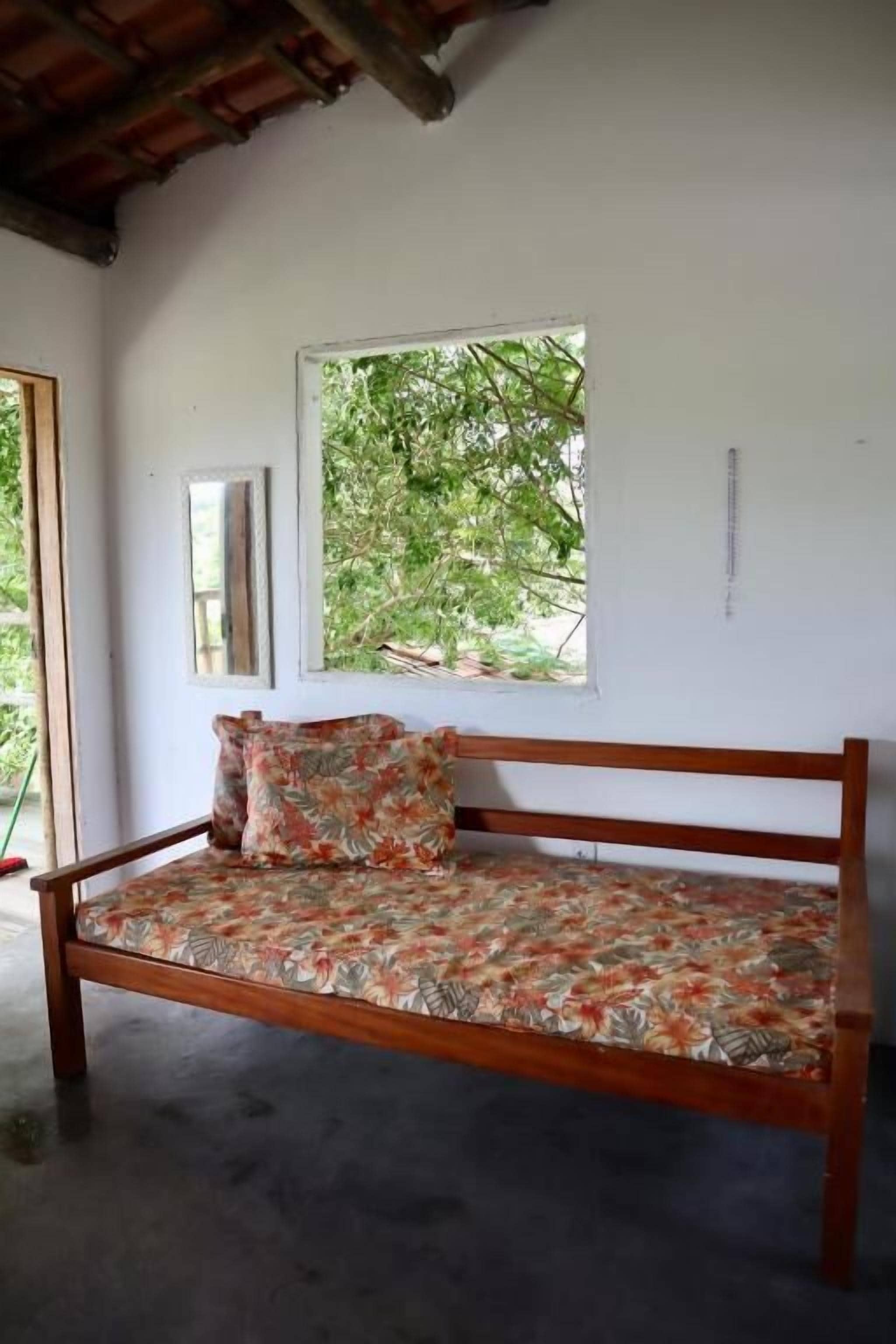 A wooden bed with a floral bedspread and pillows, a window with a view of trees, and a white wall with a mirror and a small sign.