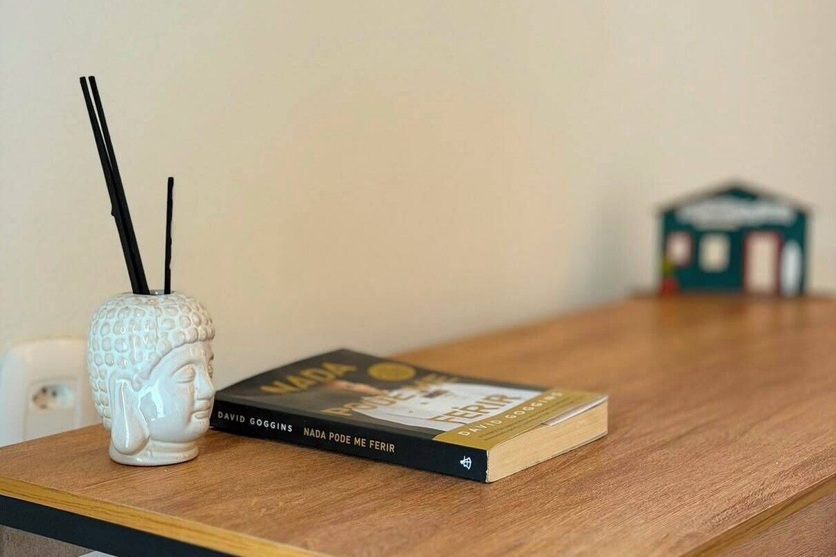 Una mesa de madera con un libro titulado 'Nada Pode Me Ferir' de David Goggins y un jarrón de cerámica blanca con detalles negros.