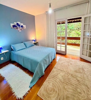 Un dormitorio con cama, mesita de noche, una obra de arte en la pared, un ventanal con cortinas y vistas a zonas verdes.