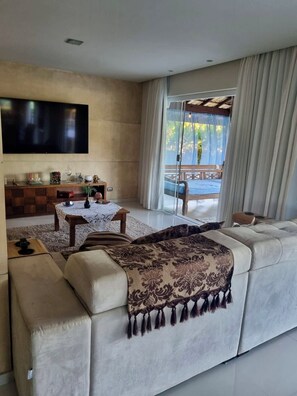 Una sala moderna con un televisor de pantalla plana grande, un sofá con un plaid estampado, una mesa de centro de madera y vistas a un balcón con mesa y sillas.
