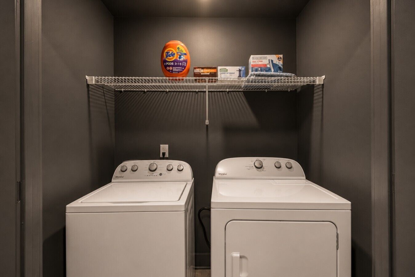 Een wasserij met twee Whirlpool wasmachines en een plank daarboven met Tide wasmiddel en andere schoonmaakproducten.