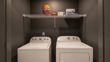 Een wasserij met twee Whirlpool wasmachines en een plank daarboven met Tide wasmiddel en andere schoonmaakproducten.