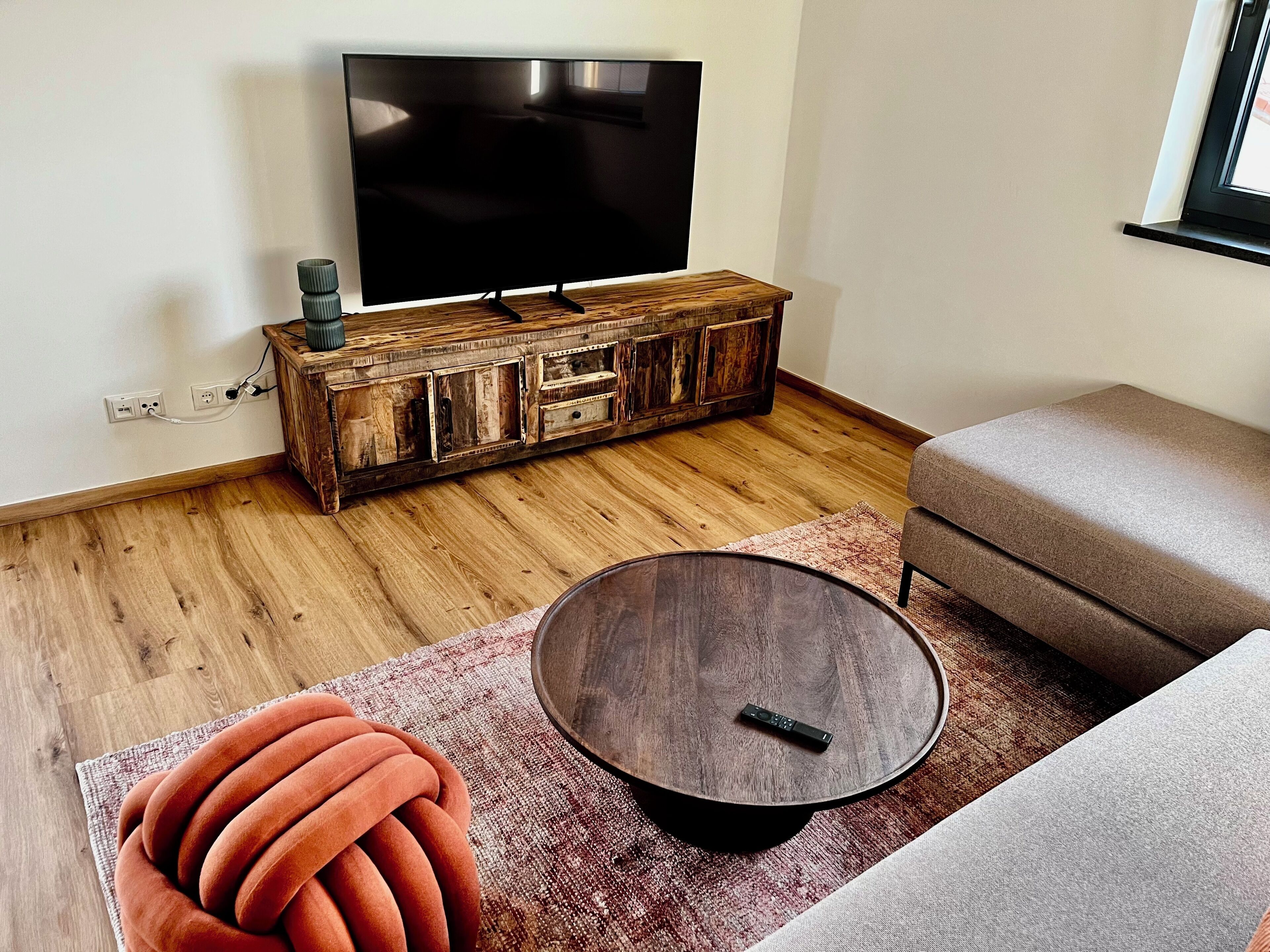 Ein Wohnzimmer mit einem Fernseher auf einer Holzkommode, einem runden Couchtisch, einem roten Sofa und einem gemusterten Teppich.