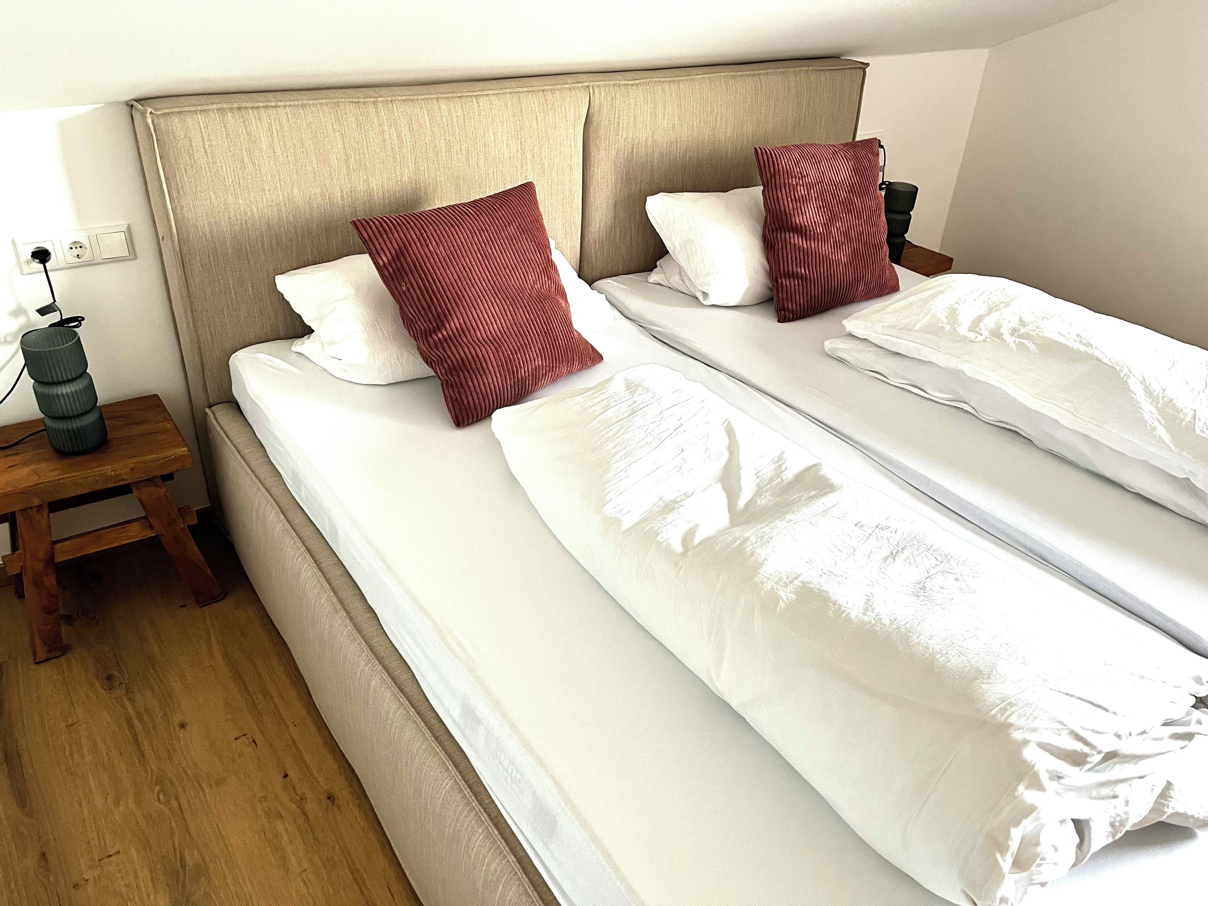 A hotel room with two single beds, each with white bedding and red pillows. There is a wooden nightstand with a lamp and a power outlet.
