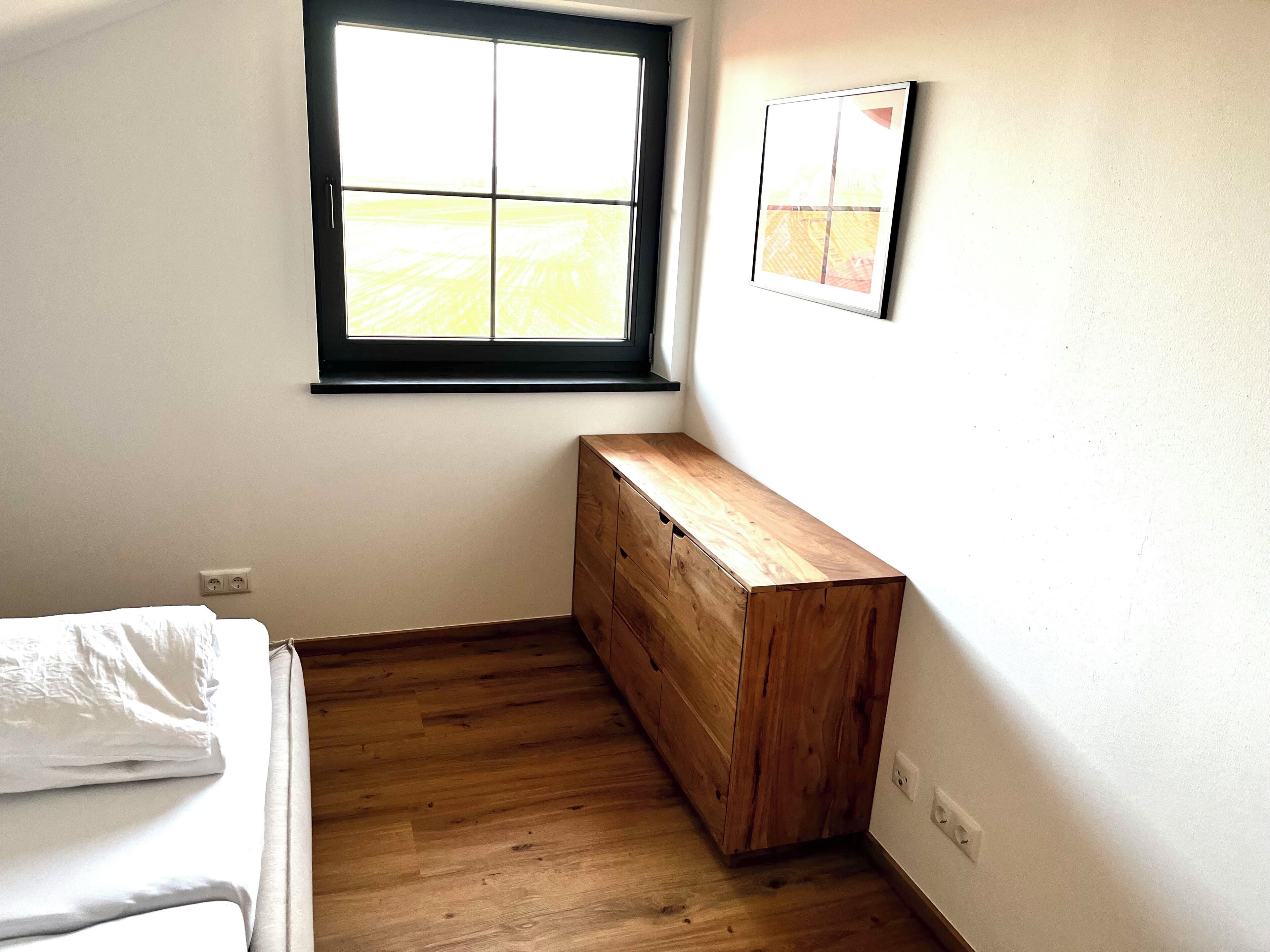 A room with a bed, a wooden bench, a window with a view, and a wall-mounted picture.