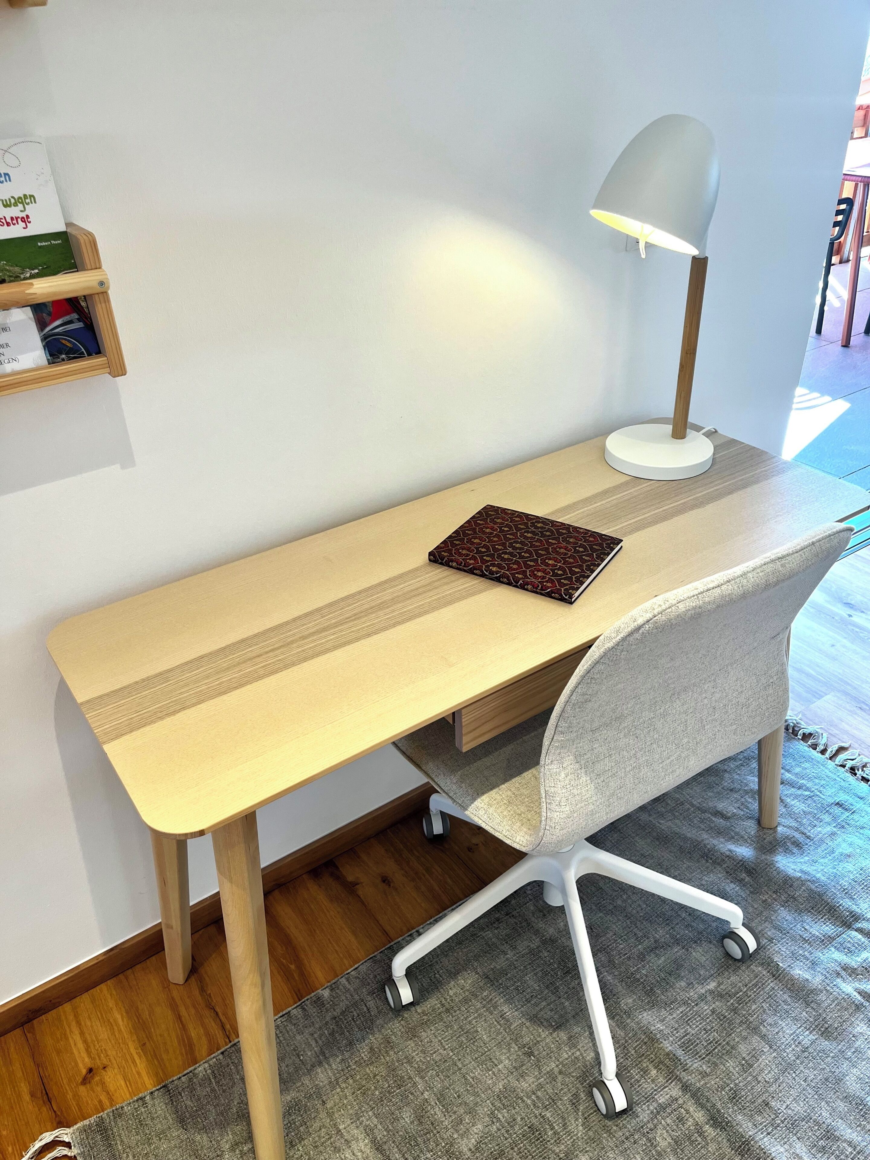 A wooden desk with a lamp, a chair, and a book on a shelf.