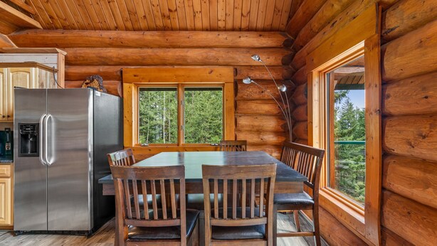 Interior de una cabaña de madera con una mesa de comedor, sillas, un refrigerador y una ventana con vistas a un bosque.