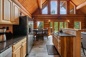 A log cabin kitchen with a central island, wooden cabinets, a stainless steel refrigerator, and a dining area with a table and chairs. - Mogul Hill Cabin - NEW Sundance Spa (Sandpoint)