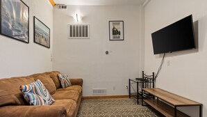 Una sala de estar con un sofá marrón, un tapete estampado, un televisor de pantalla plana montado en la pared y una pequeña mesa.