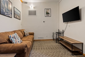 A living room with a brown sofa, a patterned rug, a flat-screen TV mounted on the wall, and a small table.