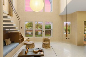 A spacious living area with a staircase, a sofa, two woven chairs, a coffee table with books and decorative items, and large windows offering views of a garden. - New listing  - Stylish Palmilla 3-Bedroom Condo with Shared Pool (Palmilla)