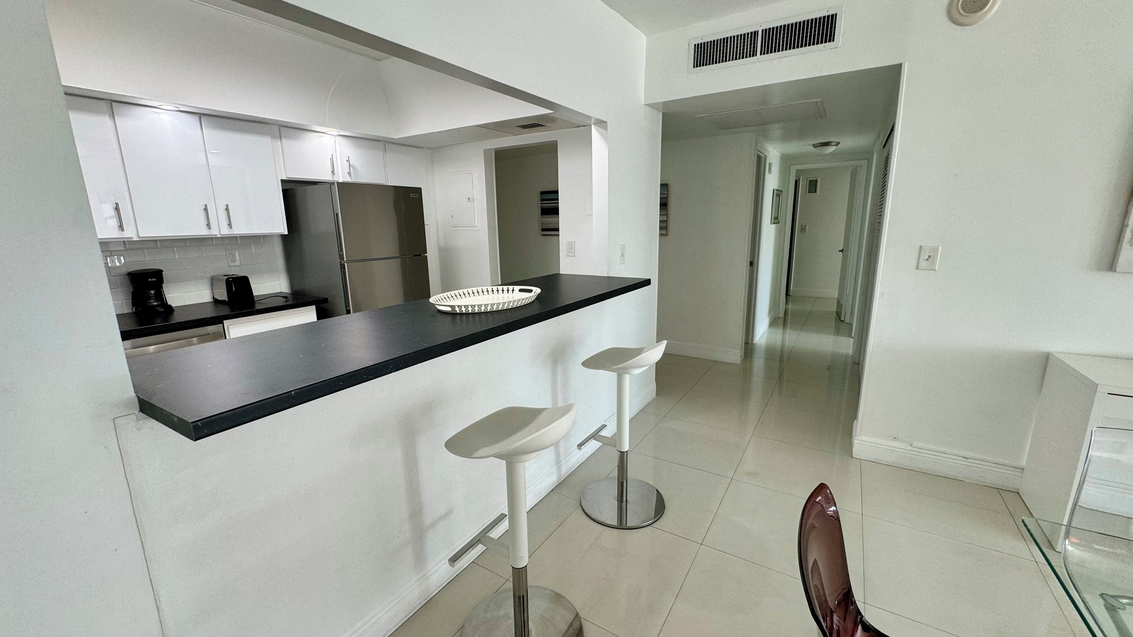 A modern kitchen with white cabinets, a black countertop, and a stainless steel refrigerator. There are two white bar stools and a glass table.