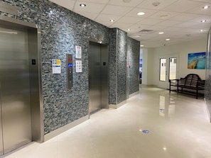 A modern elevator with a digital control panel, a tiled wall, and a bench in a clean, well-lit hallway. - Wonderful Apt! 14 floor. Parking included! (Sunny Isles Beach)