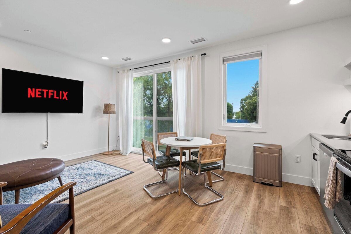 Una sala moderna con un televisor de pantalla plana que muestra 'NETFLIX', una mesa de comedor con sillas, un sofá, una pequeña cocina y un ventanal con cortinas.