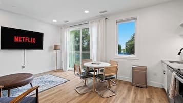 Una sala moderna con un televisor de pantalla plana que muestra 'NETFLIX', una mesa de comedor con sillas, un sofá, una pequeña cocina y un ventanal con cortinas.