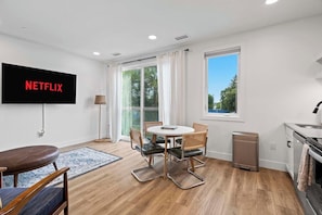 A modern living room with a flat-screen TV displaying 'NETFLIX,' a dining table with chairs, a sofa, a small kitchen area, and a large window with curtains. - West Bay Views | Dog Friendly + Near Downtown TC (Traverse City)