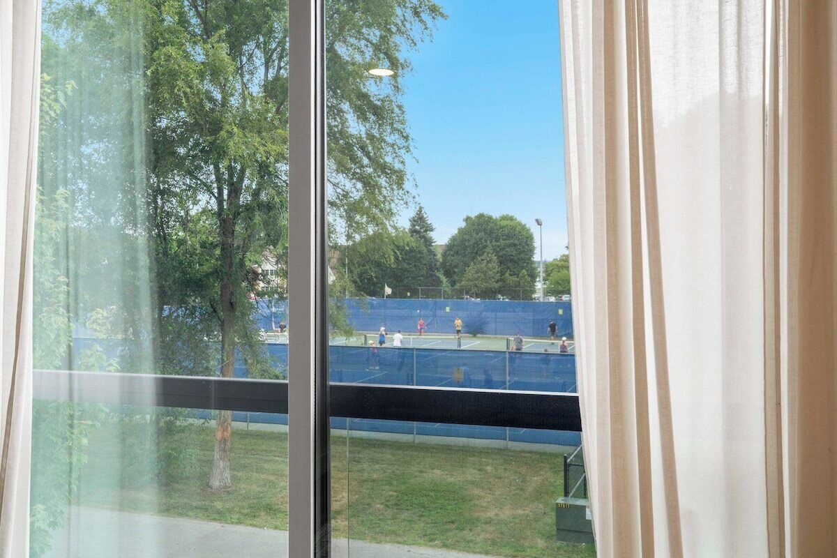Vista a través de una ventana con cortinas, que muestra una cancha de tenis y árboles al exterior.