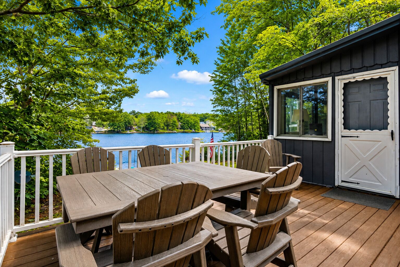 Eine Holzterrasse mit Tisch und Stühlen, mit Blick auf einen See und Häuser sowie Bäume.