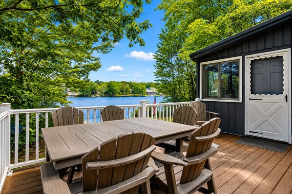 A wooden deck with a table and chairs, overlooking a lake with a view of houses and trees.