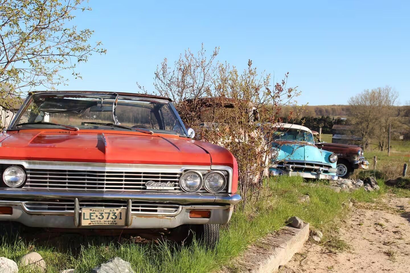 A vintage red car parked on a dirt path with grass and trees in the background.