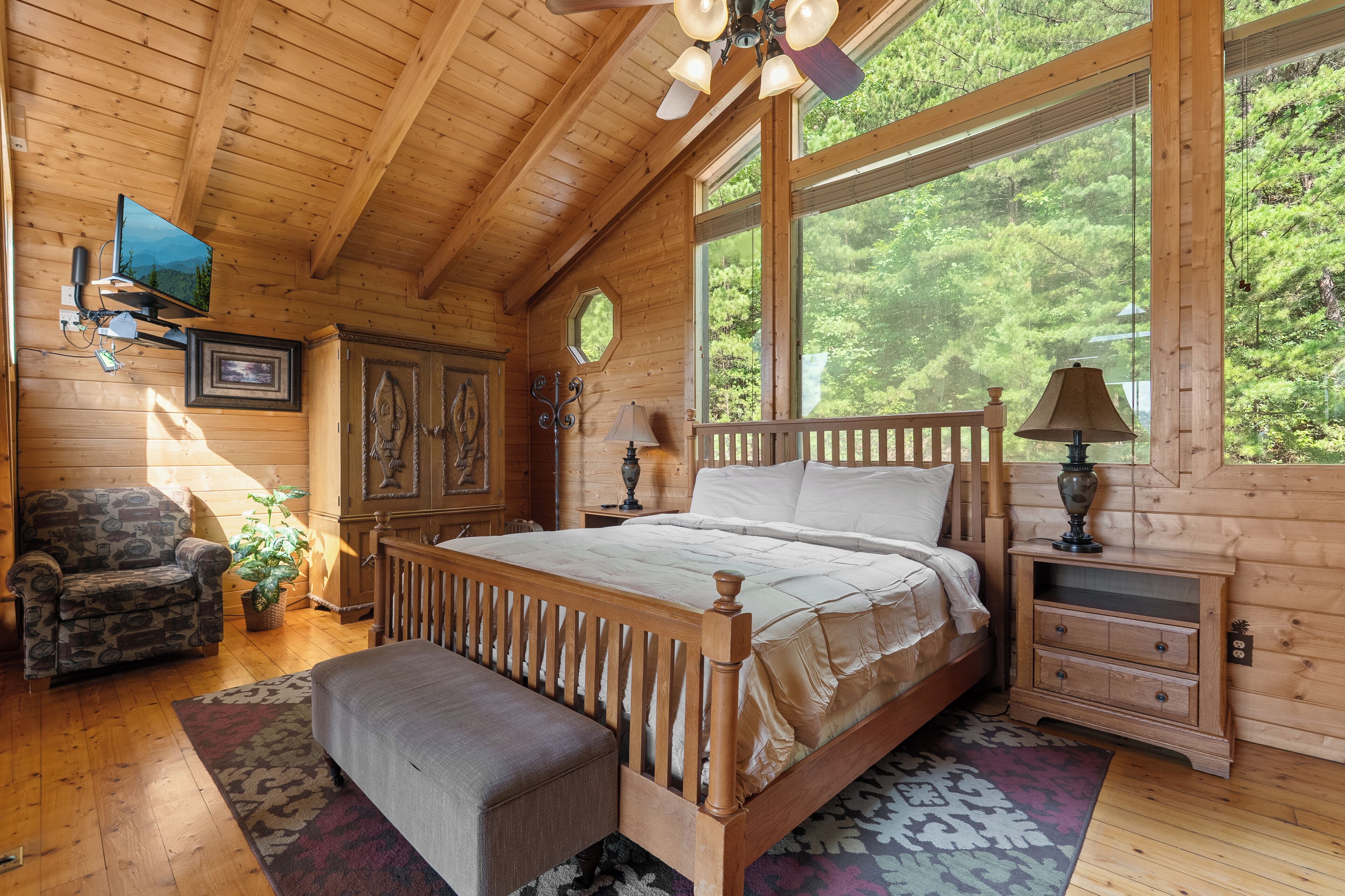 Een houten slaapkamer met een bed, een stoel, een bureau en een raam met uitzicht op groen.