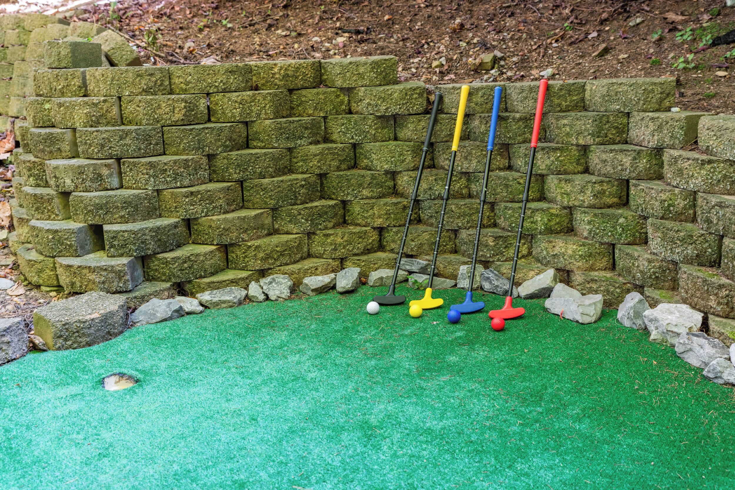 A mini-golf setup with a green artificial turf, a brick wall, and four golf clubs.