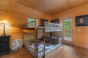 Ein Stockbett mit einer Leiter, ein Schreibtisch mit einer Lampe und ein Ventilator in einem Zimmer mit Holzwänden und einem Fenster.