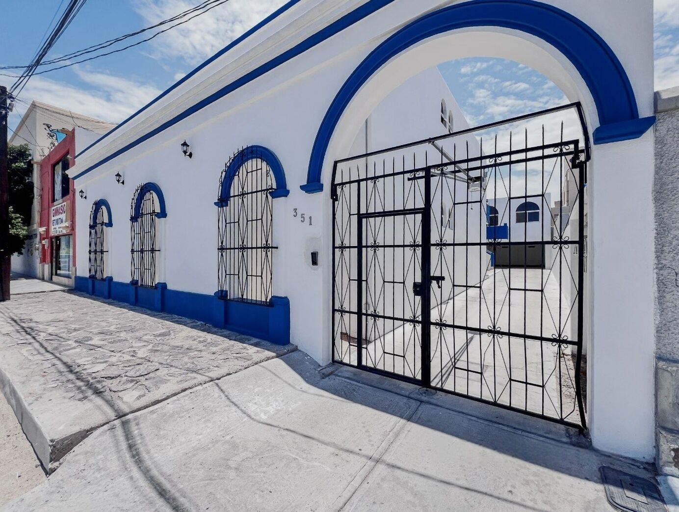 Rakennuksessa on valkoinen julkisivu ja siniset yksityiskohdat, ja siinä on suuri metallinen portti sekä kaarevat ikkunat.