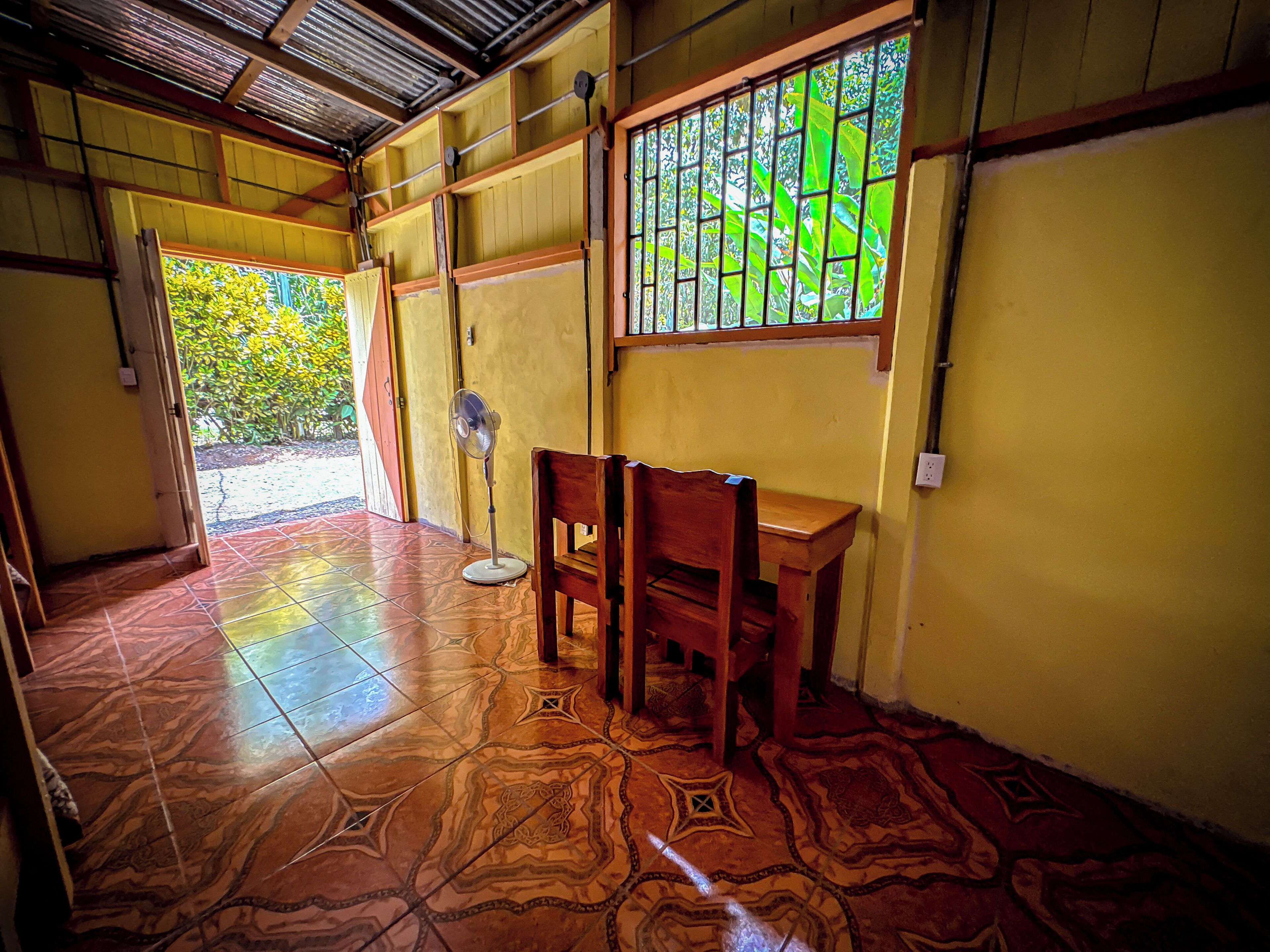 A room with a tiled floor, a wooden table, a chair, a standing fan, and a window with a view of greenery.