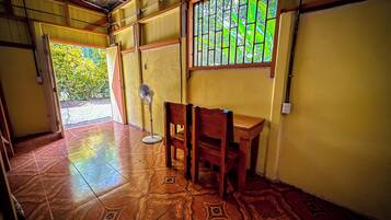 A room with a tiled floor, a wooden table, a chair, a standing fan, and a window with a view of greenery.