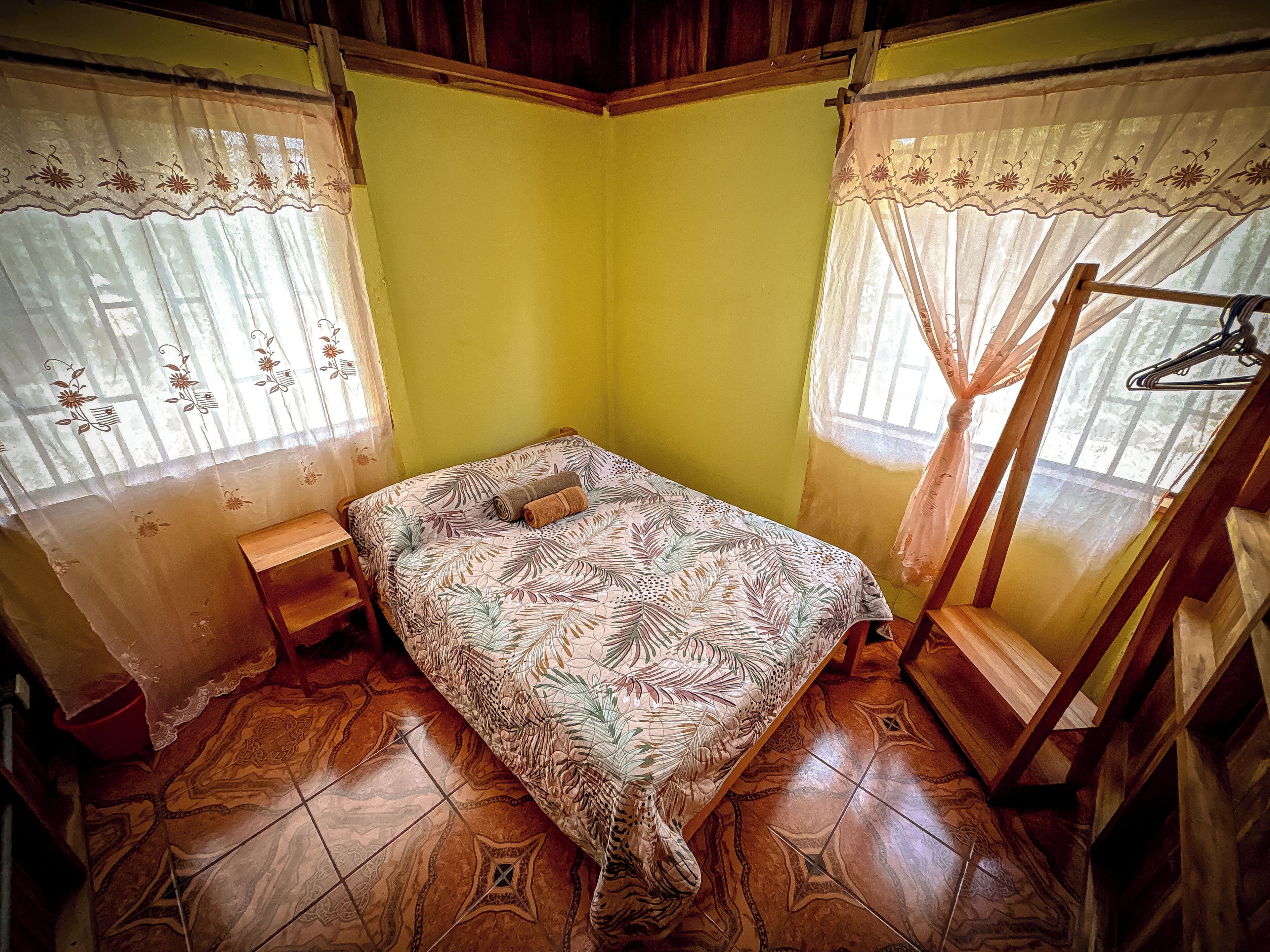 A small, single-bed room with a patterned bedspread, a wooden bench, and a mosquito net.