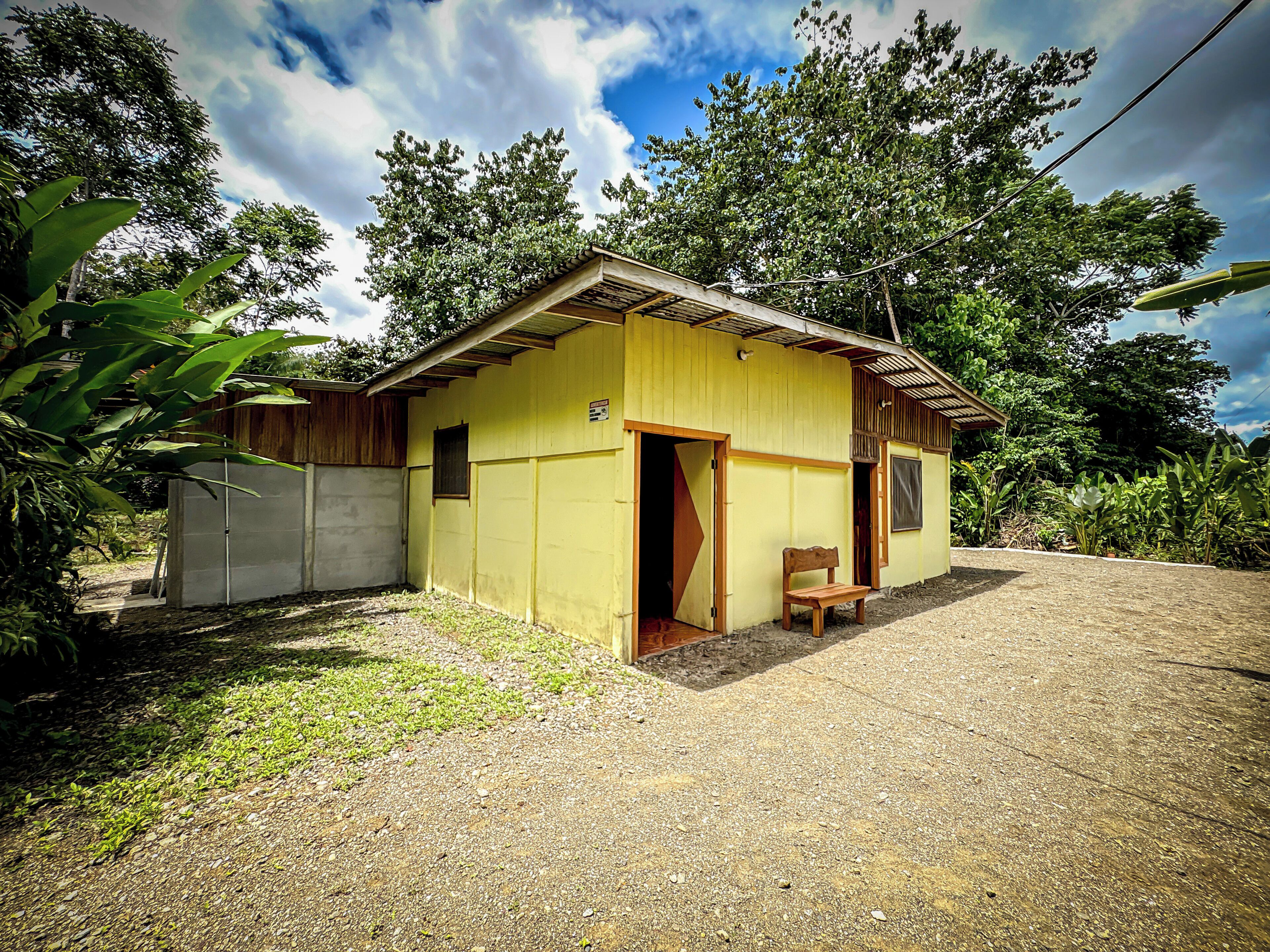 Un petit bâtiment jaune avec un banc en bois à l’extérieur, entouré de verdure.