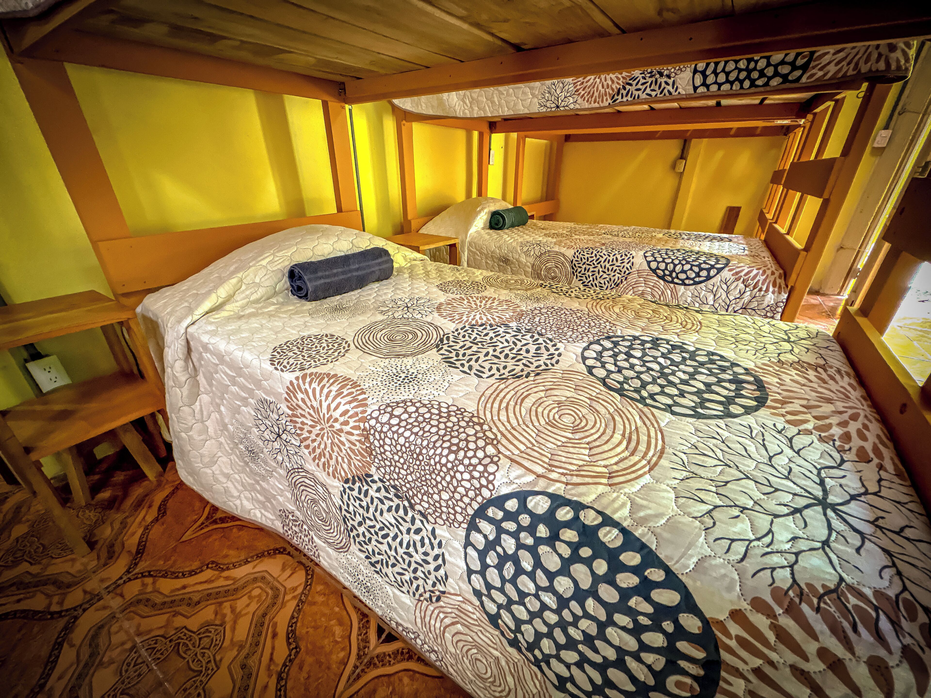 Habitación con dos literas, cada una con un edredón estampado. Se observa un banco de madera y una mesita con una lámpara.