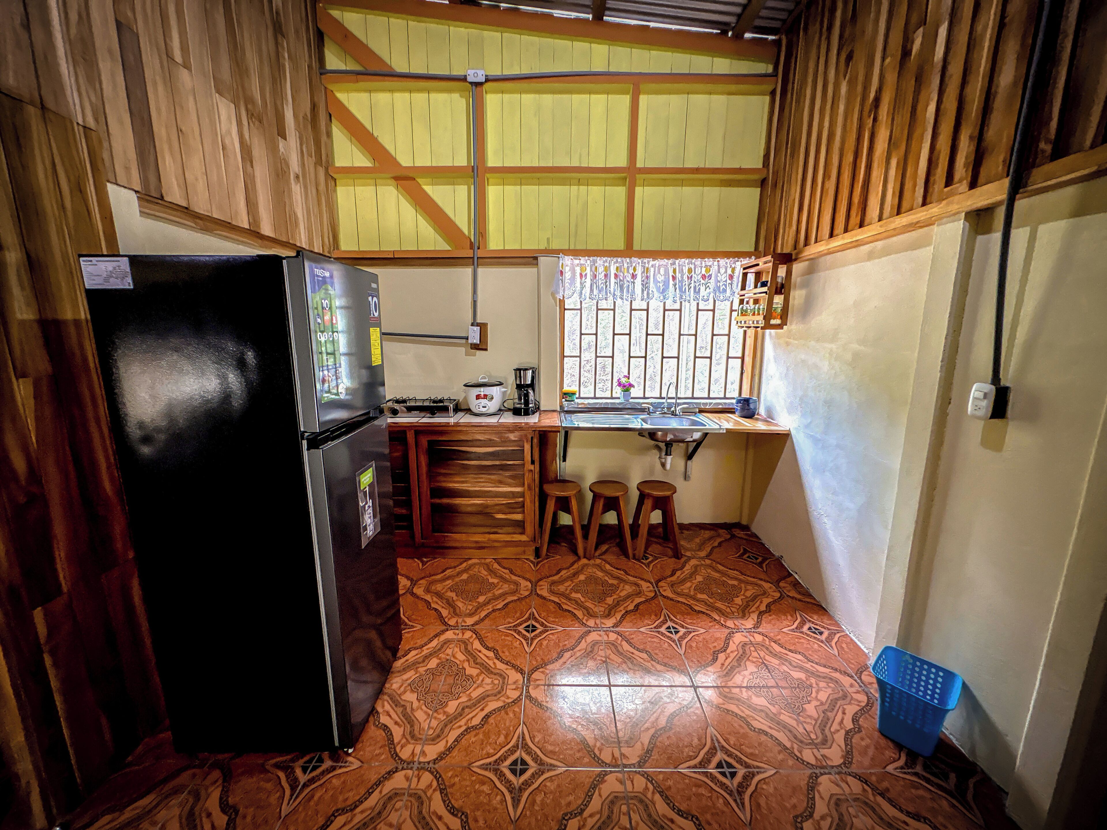 Cocina compacta con refrigerador, estufa y pileta. Hay taburetes y una ventana con cortinas estampadas.