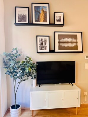 A living room with a flat-screen TV on a white cabinet, framed pictures on the wall, and a potted plant. - Vienna Garden Suites | Imperial | Terrace & Air Conditioning (Wien)