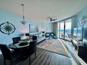 A modern living room with a dining area, a large window with a city view, and a ceiling fan. - Ocean View, 8th Floor Condo! No Hidden Fees! (Destin)