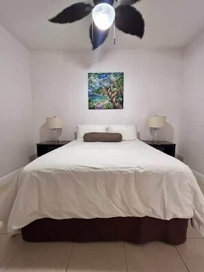 A bedroom with a bed, two bedside tables, a ceiling fan, and a wall-mounted painting. - Sunset 6 Vacations Ground Floor (Coco)