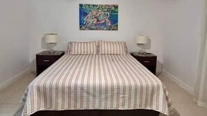 Un dormitori amb un llit, un ventilador de sostre, una taula de nit, una làmpada i un quadre a la paret.