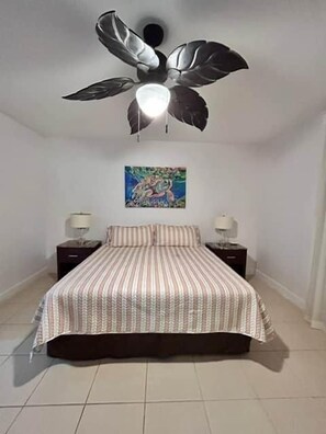 Ein Schlafzimmer mit Bett, Deckenventilator, Nachttisch, Lampe und einem Bild an der Wand.
