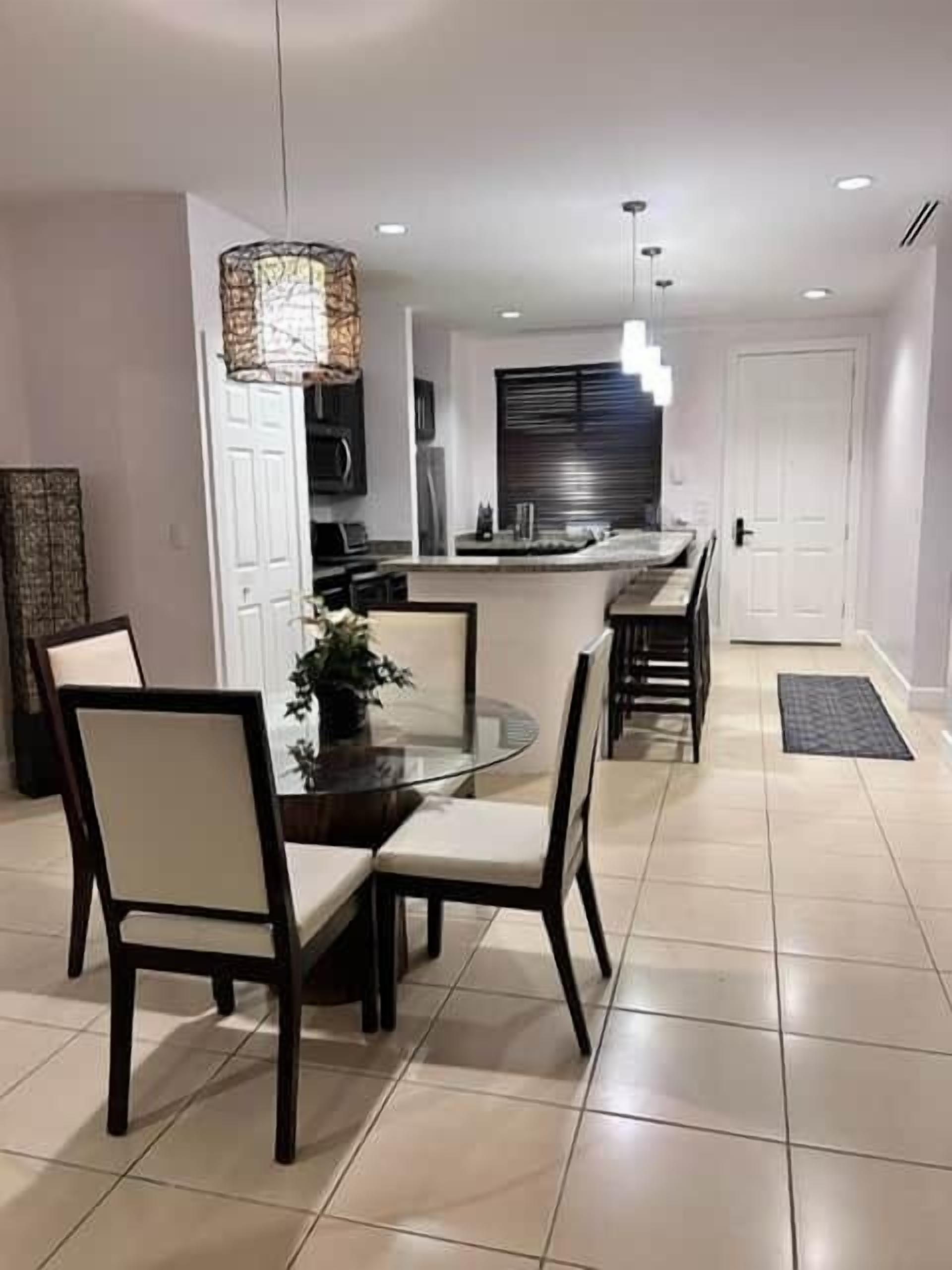 A modern dining area with a round glass table and chairs, a hanging light fixture, and a kitchen in the background.