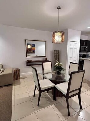 A dining area with a glass table, chairs, a hanging light fixture, a framed picture on the wall, and a door leading to a kitchen. - Sunset 6 Vacations Ground Floor (Coco)
